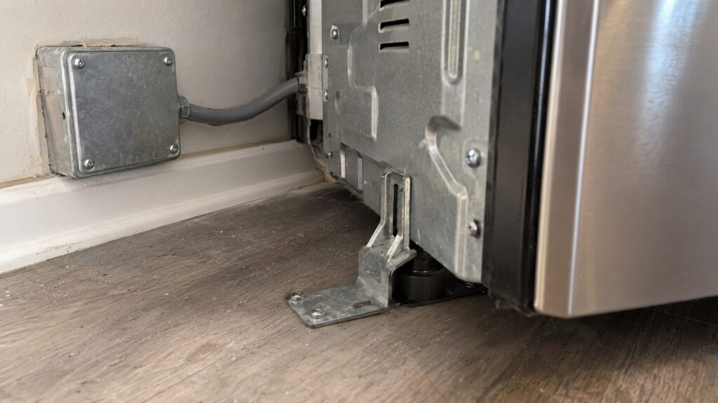 The rear lower corner of a range showing the junction box and the anti-tip bracket screwed to the floor