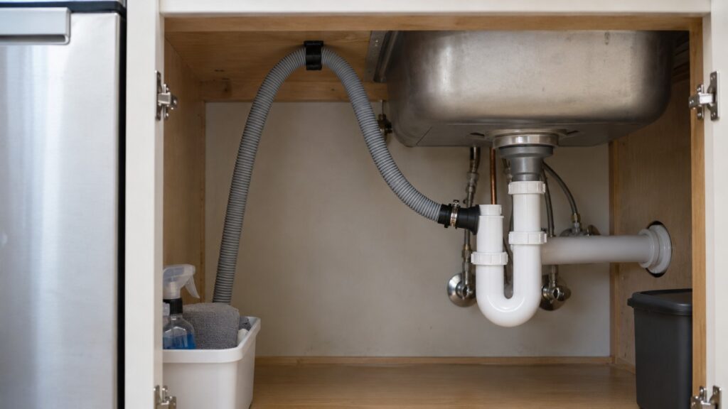 The under-sink view of a dishwasher drain hose rising into a high loop before the drain connection