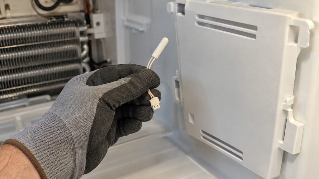 A small thermistor sensor probe held in a gloved hand next to a refrigerator evaporator cover