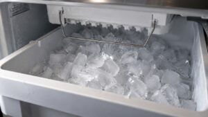 A freezer drawer interior with a mound of ice cubes heaped in the bin beneath a wire arm on an ice maker