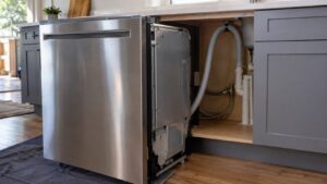 A stainless steel built-in dishwasher pulled partway out of its cabinet opening with a corrugated drain hose looped upward beneath the countertop